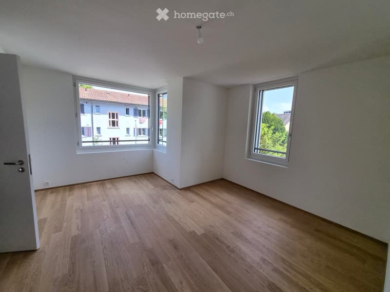 Empty room with light wooden flooring, white walls, and large windows offering views of neighboring buildings and greenery.