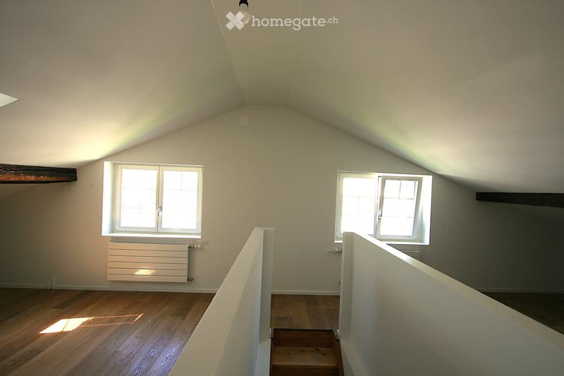 Empty attic space with wooden flooring, white walls, and two windows allowing natural light. Features a simple staircase leading upwards and a radiator on one wall.
