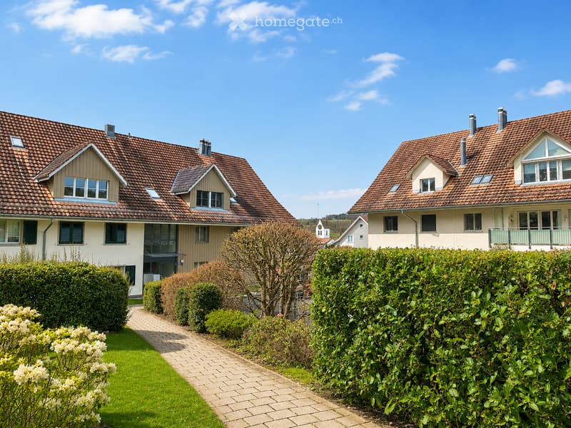 Two-story residential houses with red-tiled roofs, white walls, multiple windows, and manicured gardens with hedges and a paved pathway.