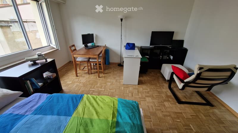 A furnished room with a bed, a chair, a table, a screen, a laptop, a shelf, and a toy. The room has wooden flooring and large windows.