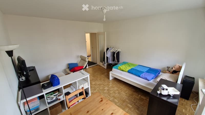 A furnished room with a bed, desk, chair, and a small table. The room features a wooden floor and white walls.