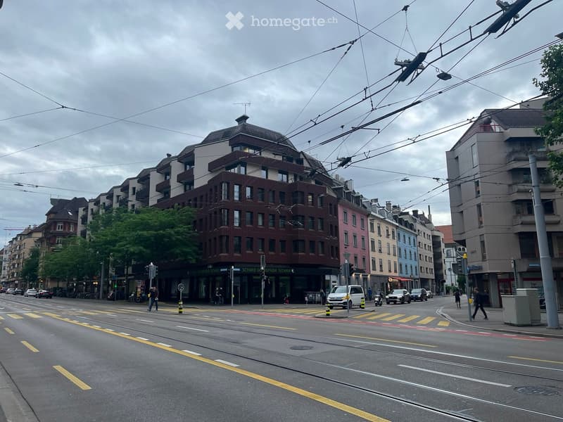 Urban street view with multi-story buildings, traffic, pedestrians, and trams. The buildings have varied facades and colors, with shops and signage visible at street level.