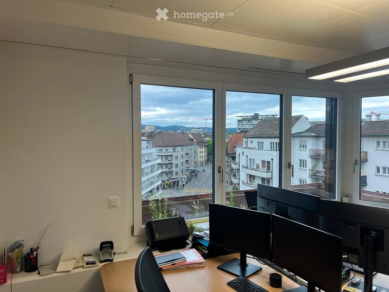 Office space with large windows overlooking a cityscape, featuring a desk with multiple monitors, a chair, a printer, and various office supplies.