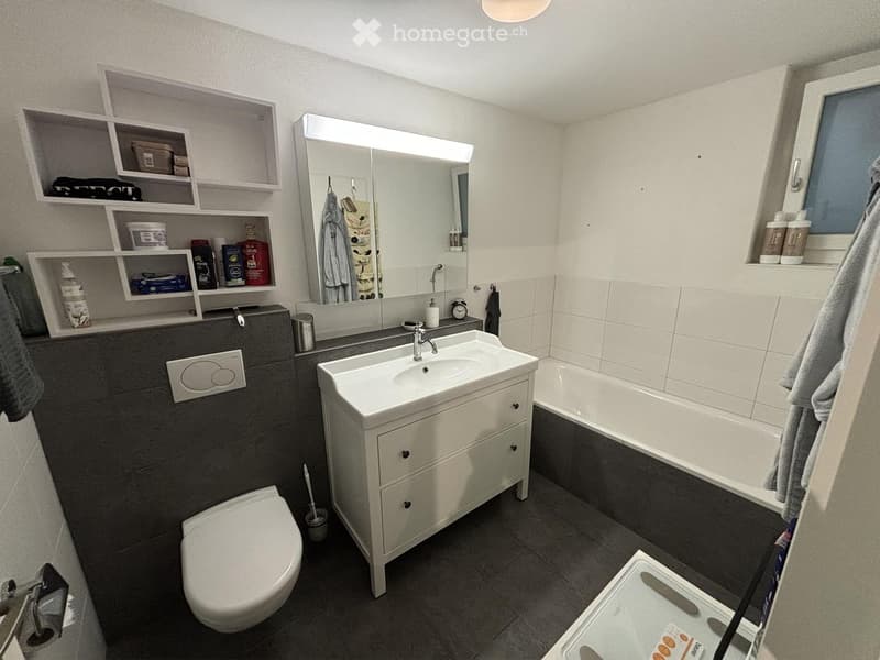 Modern bathroom with a white sink, toilet, bathtub, large mirror, and built-in shelves holding toiletries. Dark grey tiles on the floor and walls.