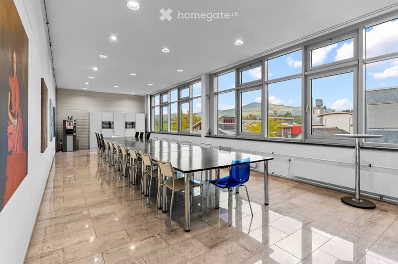 Bright, modern conference room with large windows, a long glass table surrounded by chairs, a small kitchen area with a coffee machine, and a view of the landscape.