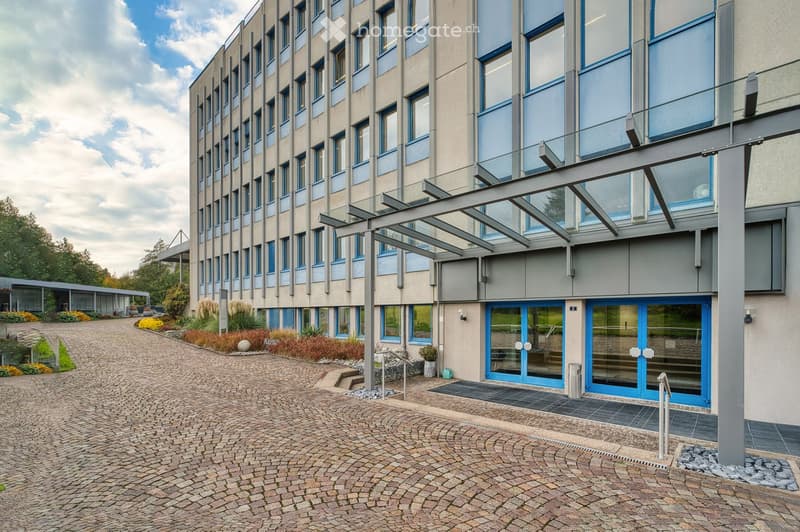 Modern multi-story building with large windows and blue accents, featuring a covered walkway and landscaped entrance area.