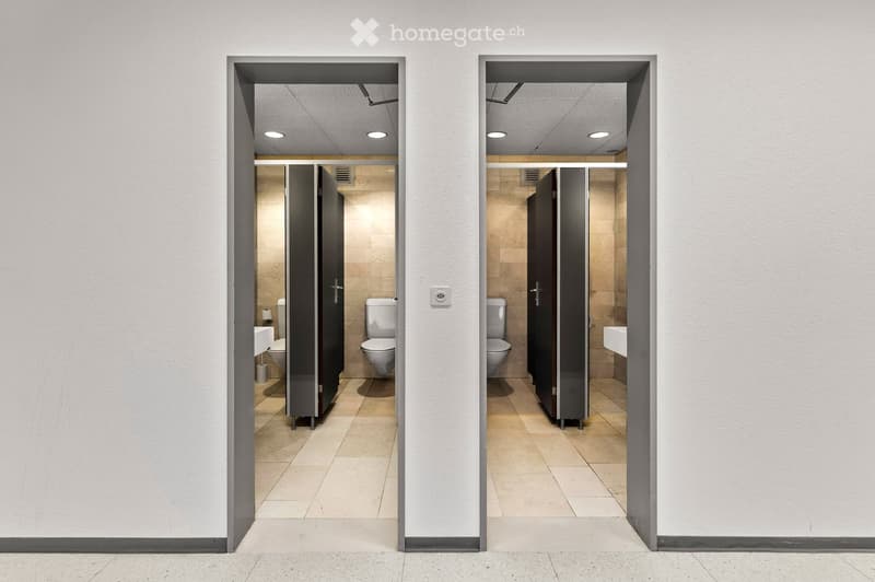 Two adjacent restrooms with tiled floors and walls, each containing a toilet and a sink, separated by stall doors.