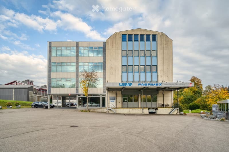Modern two-story building with large windows and a sign reading "WERAP FABRIMEX Hauptingang/Anmeldung" above the entrance. The building has a concrete facade and is surrounded by a paved area with several parked cars.