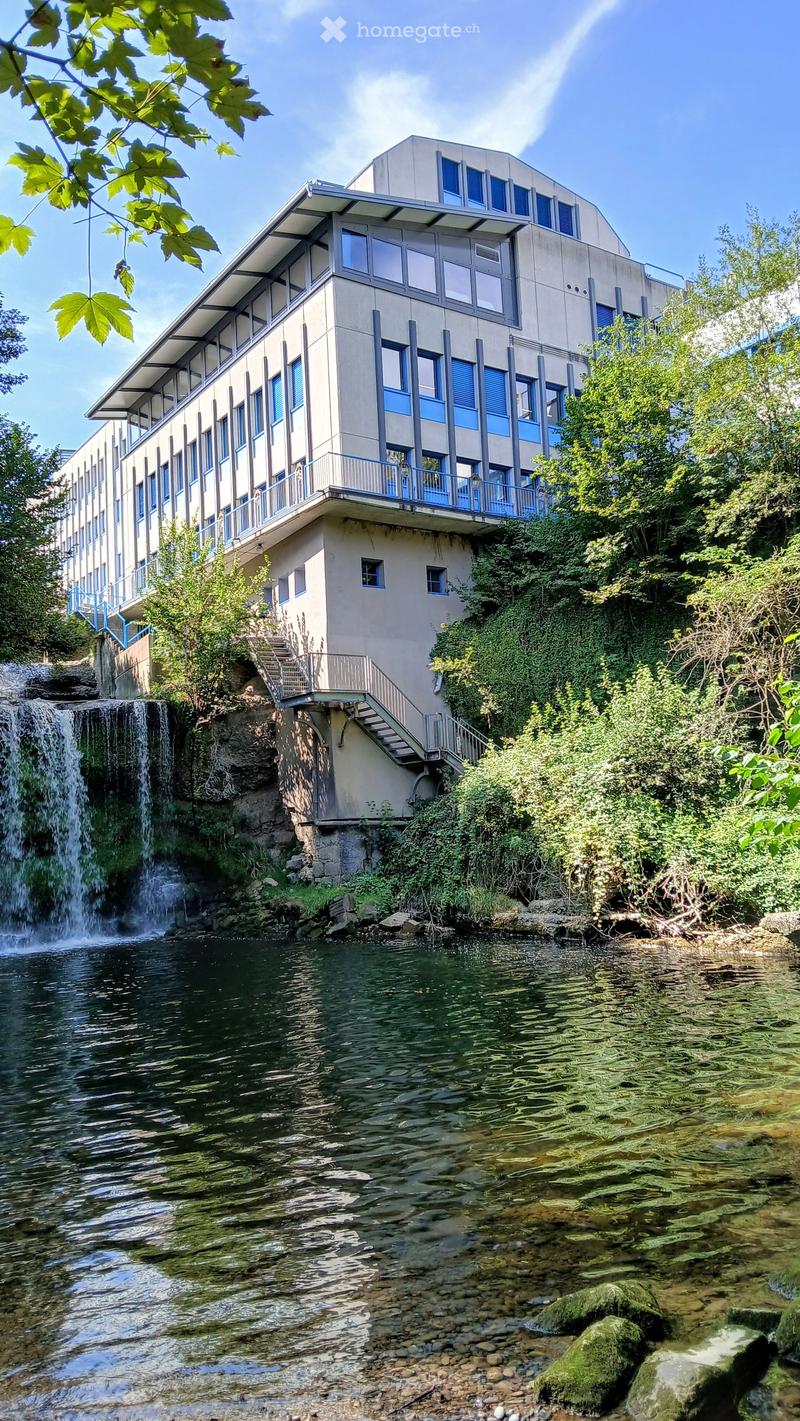 Modern building with multiple floors, blue railings, large windows, balcony, located on a hill with a waterfall