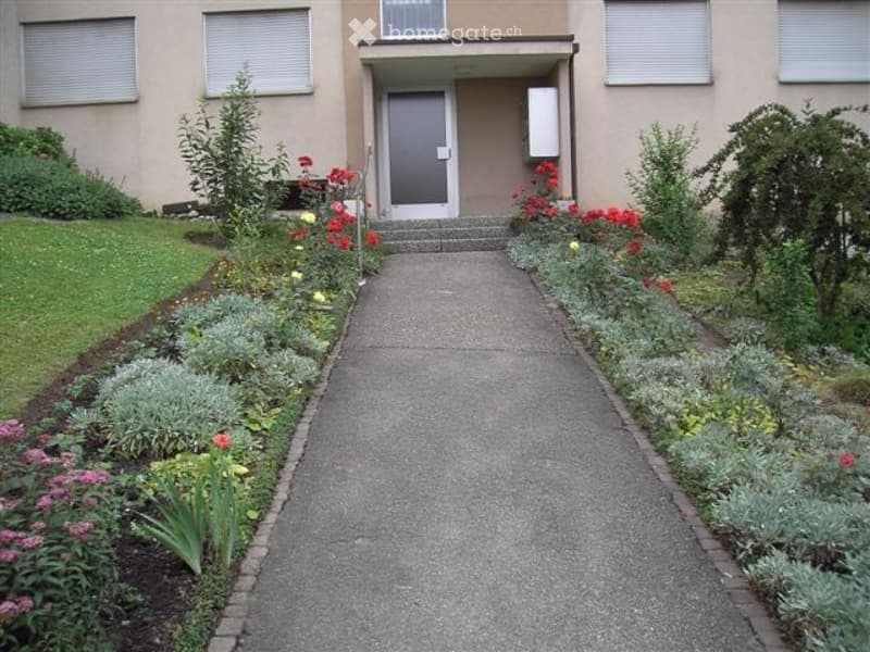 A well-maintained garden path leads to the entrance of a building, flanked by neatly arranged flower beds and shrubs.