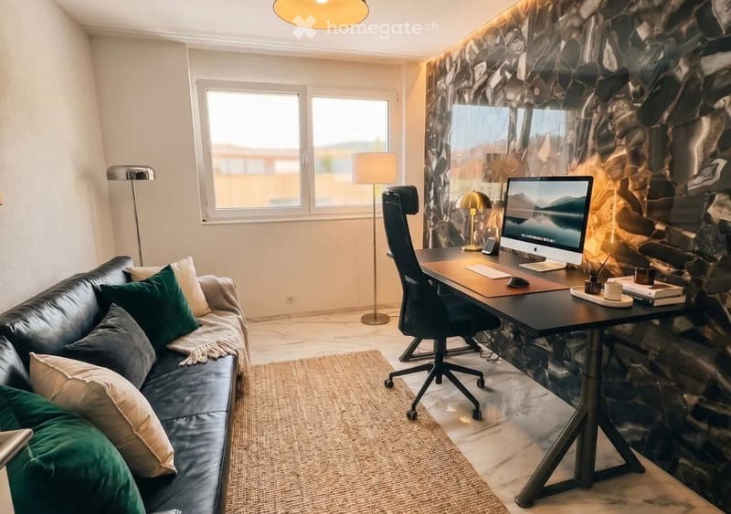 A modern, well-lit room with a black leather sofa, a wooden desk with a computer, a chair, and a lamp. The room features large windows, a stone accent wall, and a round rug on the floor.