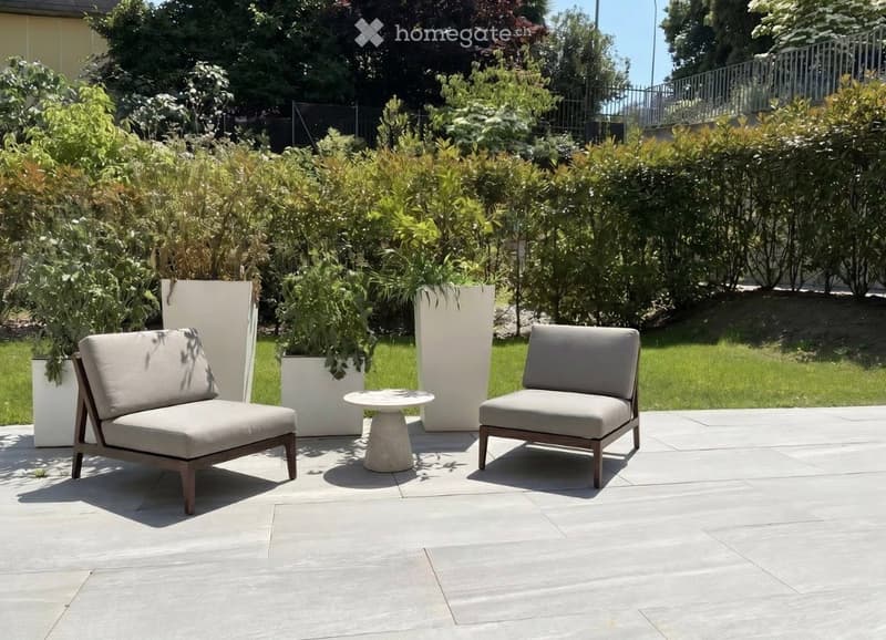A modern outdoor seating area with two beige chairs and a small round table on a concrete patio, surrounded by lush greenery and tall white planters.