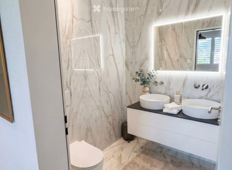 Modern bathroom with marble walls and floors, double sink vanity with modern faucets, large mirror with LED lighting, and a toilet visible in the background.