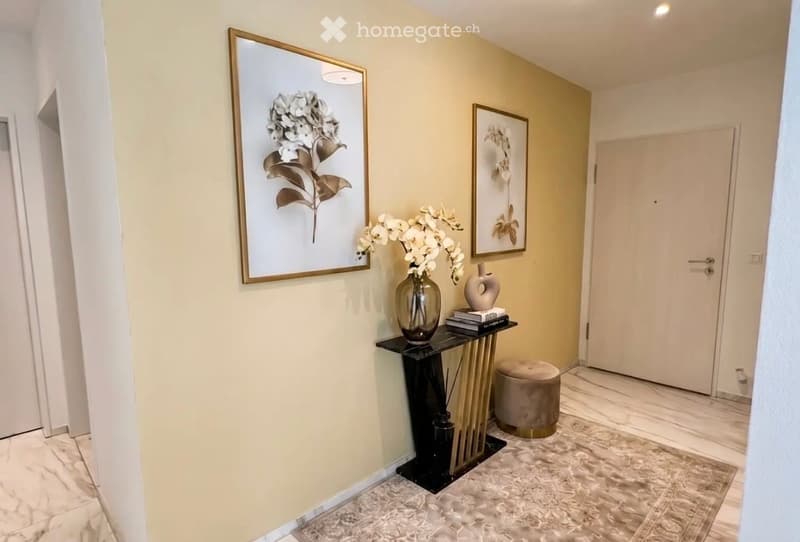 Elegant hallway with marble flooring, a small black console table with a vase of white flowers, a round stool, and framed artwork on the walls.