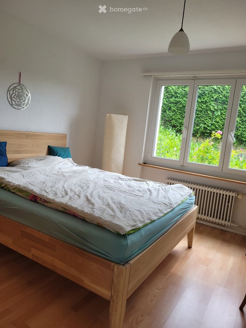 Cozy bedroom with wooden bed, white bedding, blue pillows, radiator, and wall decoration. Large windows offer a view of greenery outside.