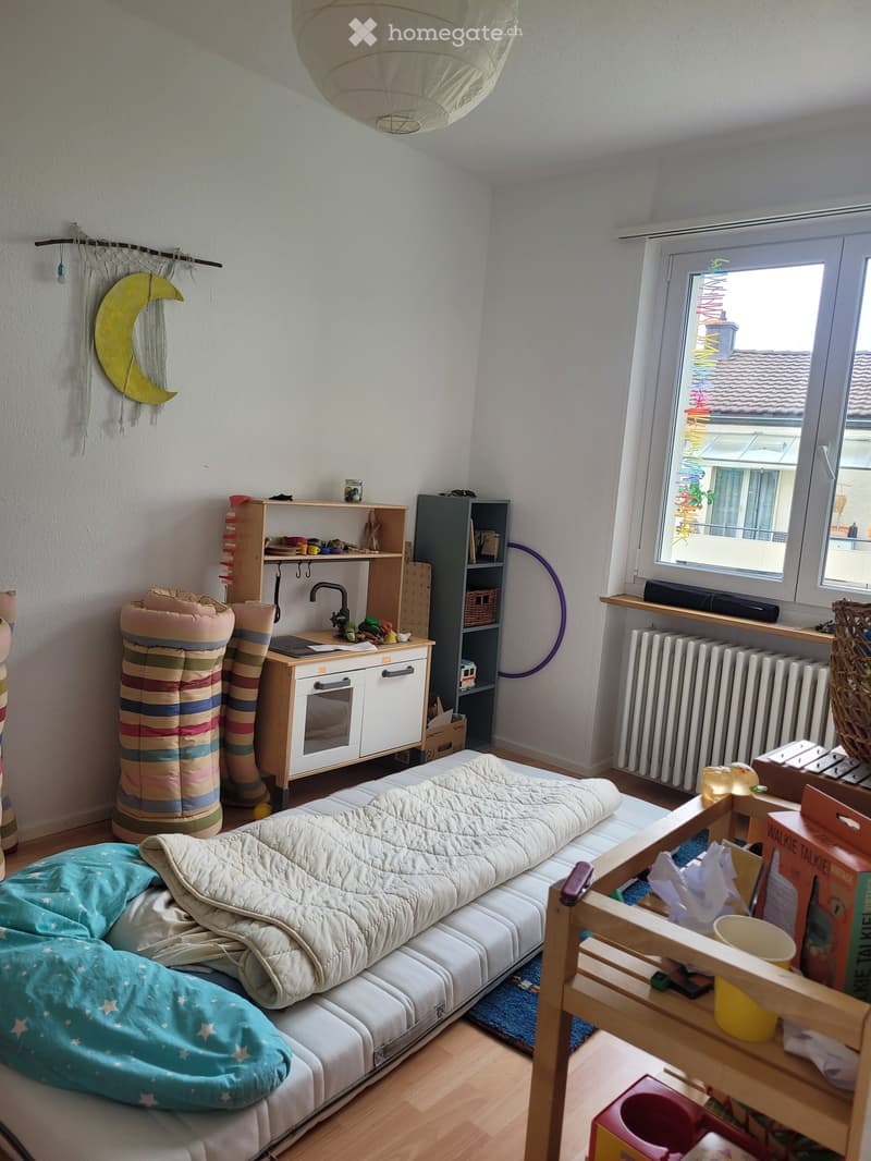 A cozy, well-lit bedroom with a sofa bed, a blonde wood cabinet, a shelf, a radiator, a table, and various toys and items.