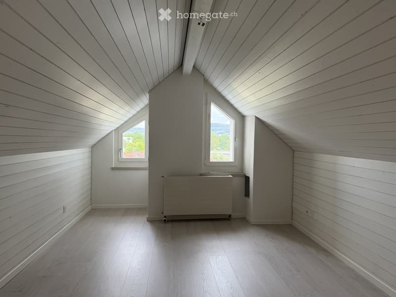 Empty attic room with wooden walls, slanted ceiling, wooden floor, and two windows offering a view of the outdoors.