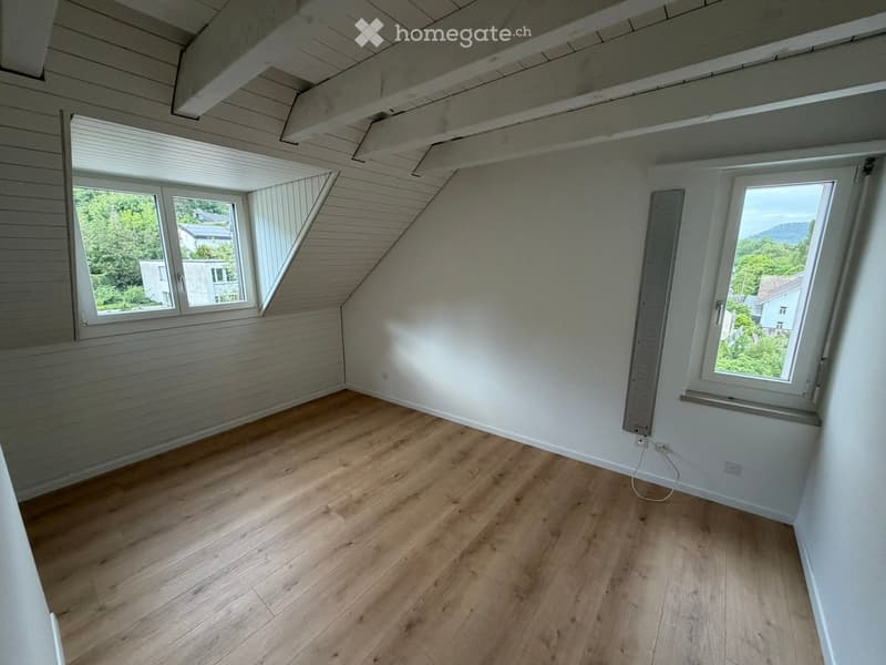 Empty attic room with wooden flooring, white walls, and exposed wooden beams on the ceiling. Two windows provide natural light and views of the surrounding area.