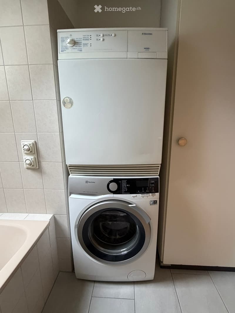 A compact bathroom with a white washing machine and a white dishwasher stacked vertically, tiled walls, and a small sink visible on the left side.