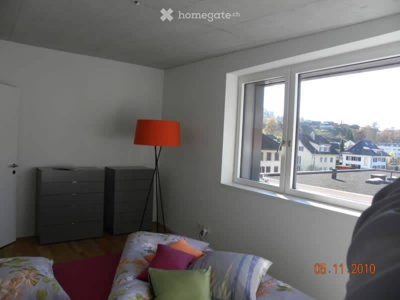 Bright bedroom with wooden flooring, large window offering a view of the city, a neatly made bed with colorful pillows, a floor lamp, and a grey drawer unit.