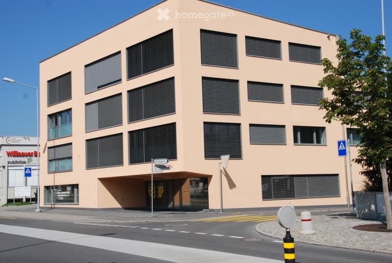 Modern, multi-story building with a light pink facade and numerous windows with blinds. The building has a clean, contemporary design with a flat roof and a small entrance area visible at the bottom.