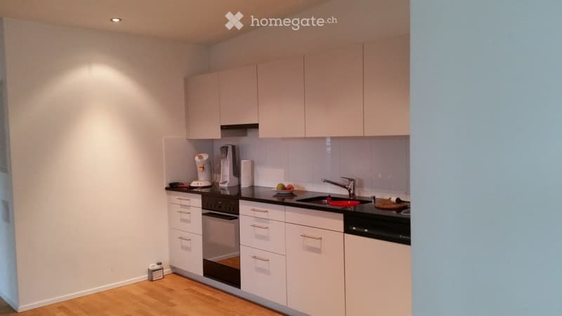 Modern kitchen with white cabinets, black countertops, and stainless steel appliances. Features include an oven, sink, faucet, fridge, coffee maker, and a small fruit bowl on the counter.