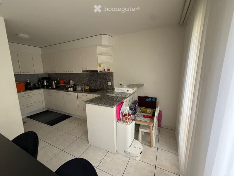 Modern kitchen with white cabinets, granite countertop, and various appliances. Includes a small dining area with a table and chair, a trash bin, and a vacuum cleaner on the floor.