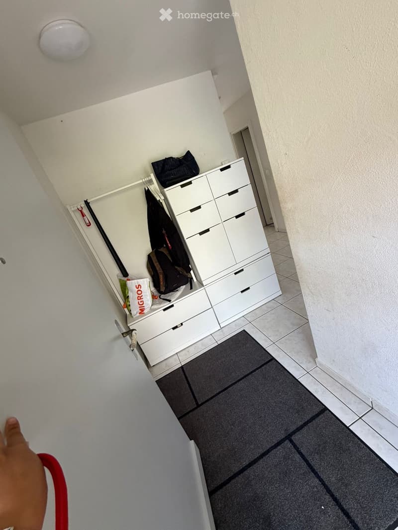 A clean, modern hallway with white walls, tiled flooring, and a built-in white storage unit with multiple drawers. A black rug lies on the floor, and a coat rack with a few items is visible.