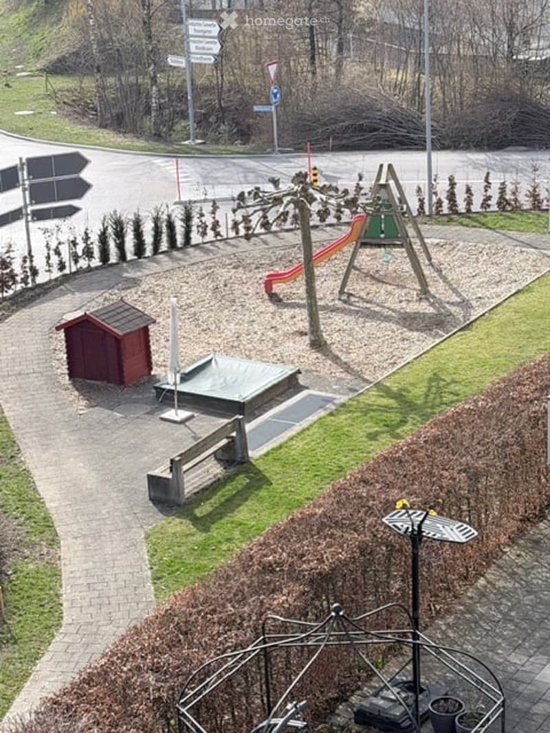 Outdoor playground area with slide, seesaw, small storage shed, paved pathway, benches, hedges, and plants.