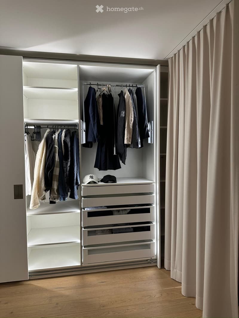 A modern, well-organized walk-in closet with white built-in wardrobe and drawers, neatly arranged clothing, and two hats on the shelf.