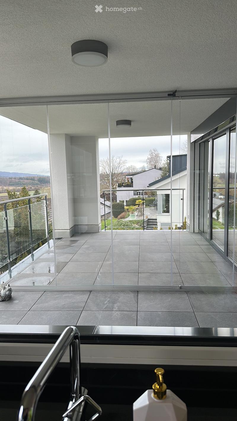 Modern balcony with large glass sliding doors, tiled floor, metal railing, and a view of a landscaped garden and neighboring house.