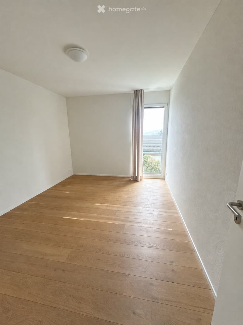 Empty room with wooden flooring, white walls, and a large window with a curtain. The room has a modern design with a visible light fixture on the ceiling and a door handle on the right side.