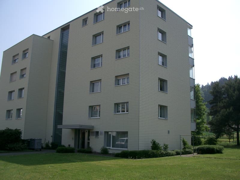 Modern multi-story building with light-colored facade, multiple windows, and a small entrance area surrounded by greenery.