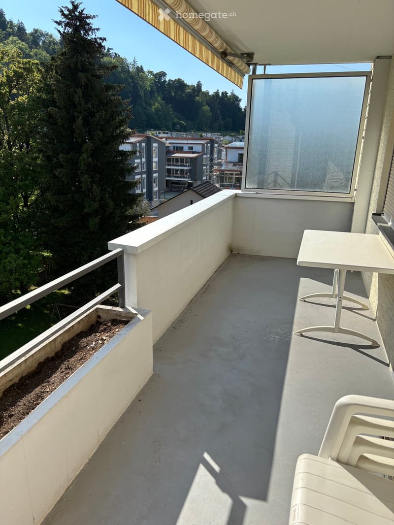 Balcony with a table and chairs, overlooking a green, tree-lined area with buildings in the distance.