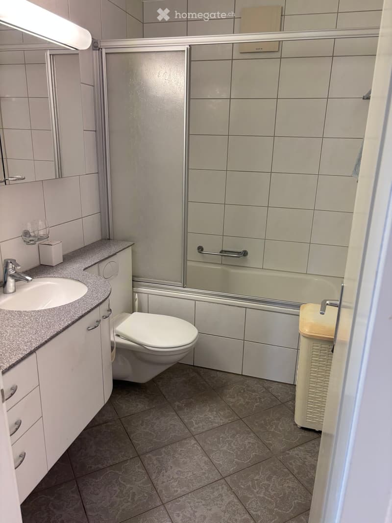 Modern bathroom with white tiles, a sink with a faucet, a toilet, a bathtub with a glass enclosure, and a small cabinet with a mirror above it.