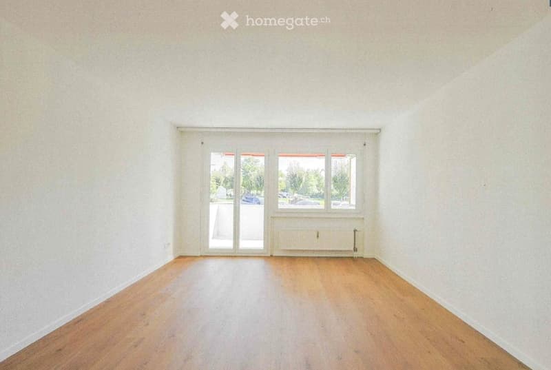 Empty room with wooden flooring, white walls, and large windows with a sliding door leading outside.