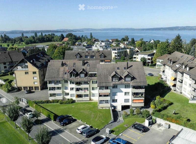 Aerial view of a multi-story residential building complex with a parking lot, surrounded by greenery and other buildings.