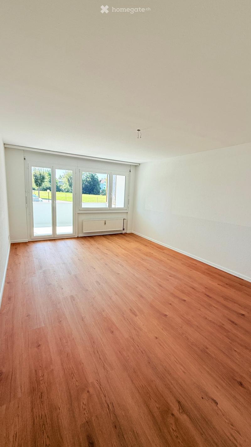 Empty room with wooden flooring, white walls, and large windows with a sliding door leading outside.