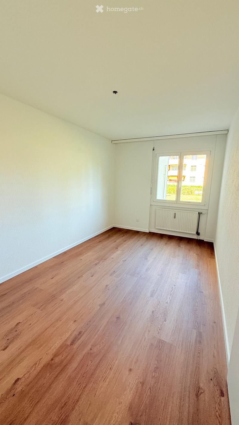 Empty room with light-colored walls and wooden flooring, featuring a window with white frames and a radiator below it.