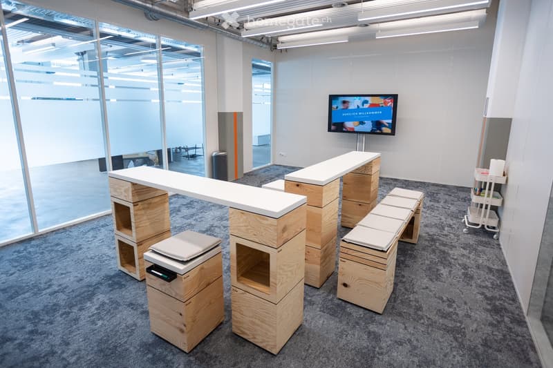 Modern reception area with a white table, wooden stools, a television screen, and a cart with supplies.