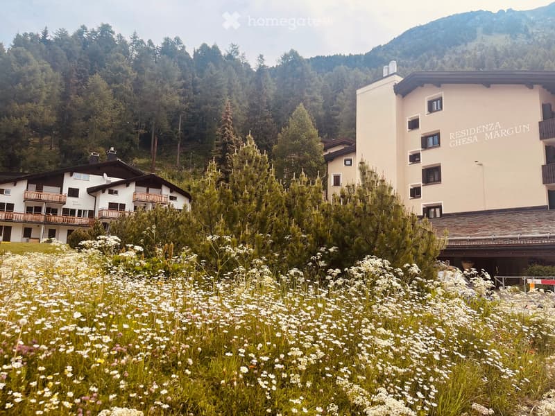 A scenic view of a residential area surrounded by lush greenery and wildflowers, with two buildings visible, one labeled "RESIDENZA CHESA MARGUN" and another with balconies.