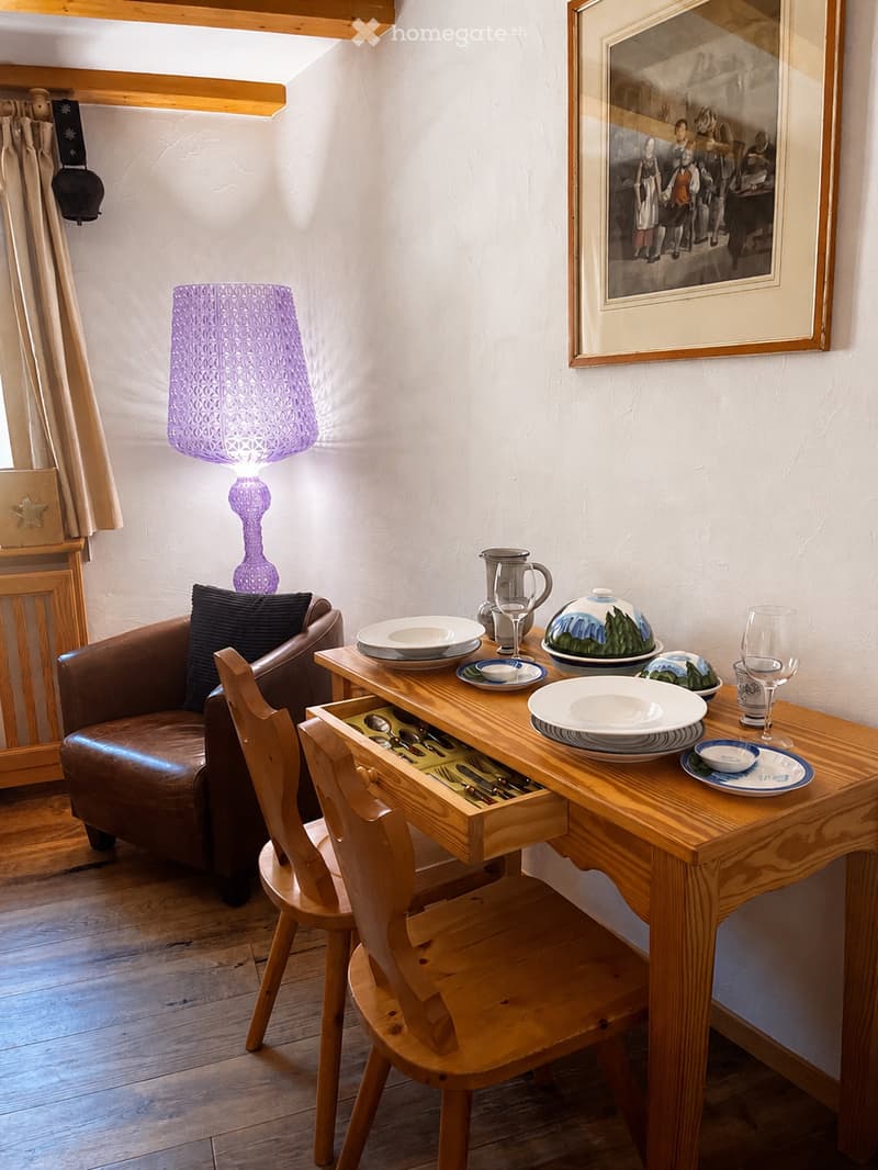 A cozy, well-furnished room with a wooden dining table set for a meal, a brown leather sofa, a wooden chair, and a decorative purple lamp. The room features a framed picture on the wall and a curtained window.