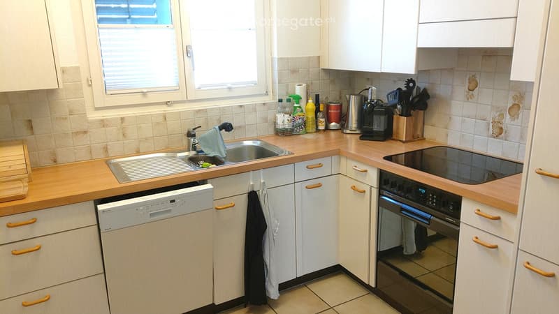 A compact kitchen with white cabinets, wooden countertops, a sink, dishwasher, and oven. Various kitchen utensils and bottles are placed on the counter.