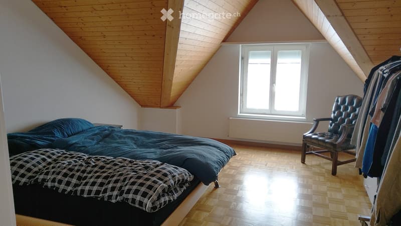 Bright attic bedroom with wooden slanted ceiling, large window, bed with dark blue bedding, wooden chair, and clothes hanging on a rack.