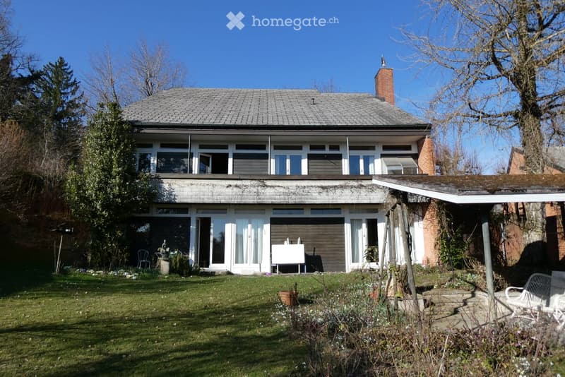House. Two-story house with a flat roof, large windows, and a covered patio area. The house features a well-maintained garden with a lawn, potted plants, and outdoor seating.