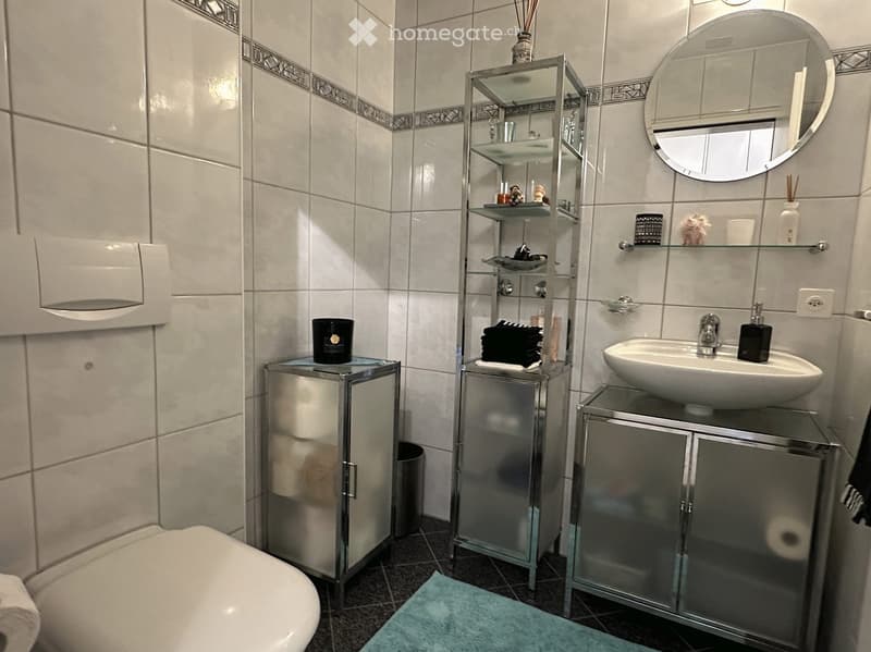 white tiled walls, white toilet, white sink, glass shelves, round mirror, blue rug