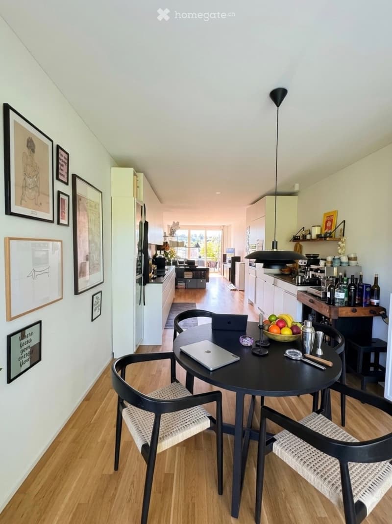 A modern, open-plan kitchen and dining area with light-colored cabinets, a black dining table with four chairs, and various kitchen appliances visible in the background.