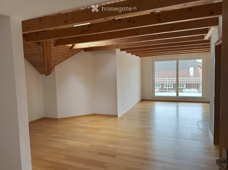 Empty room with wooden flooring and exposed wooden beams on the ceiling. Large window with a view of the outside.