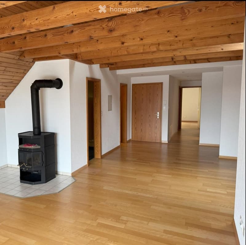 Empty hallway with wooden flooring, white walls, and exposed wooden beams on the ceiling. A black wood-burning stove is visible on the left side.