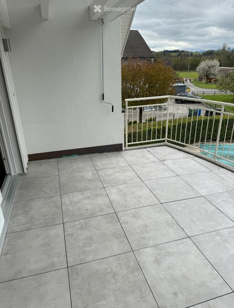 Balcony with tiled floor, white railing, and a view of a residential area with trees and a swimming pool.